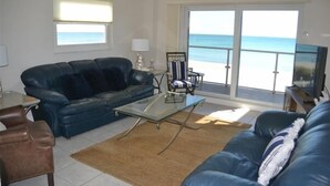 A living room with a blue sofa, a glass coffee table, a brown rug, and a view of the beach through large glass doors. - Beachcomber unit 202 (Longboat Key)