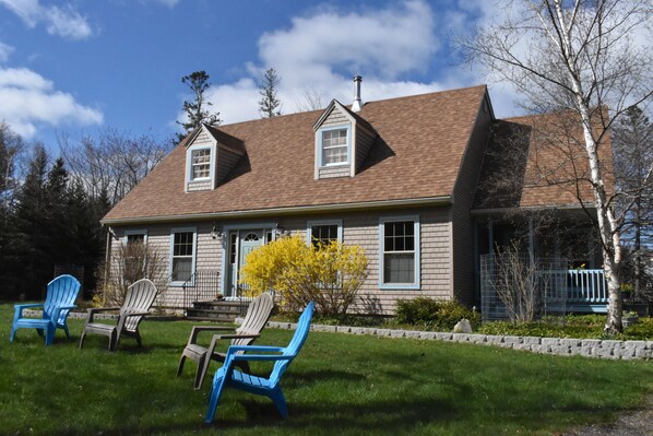 A two-story house with a brown roof, a front porch, and a small garden with chairs.