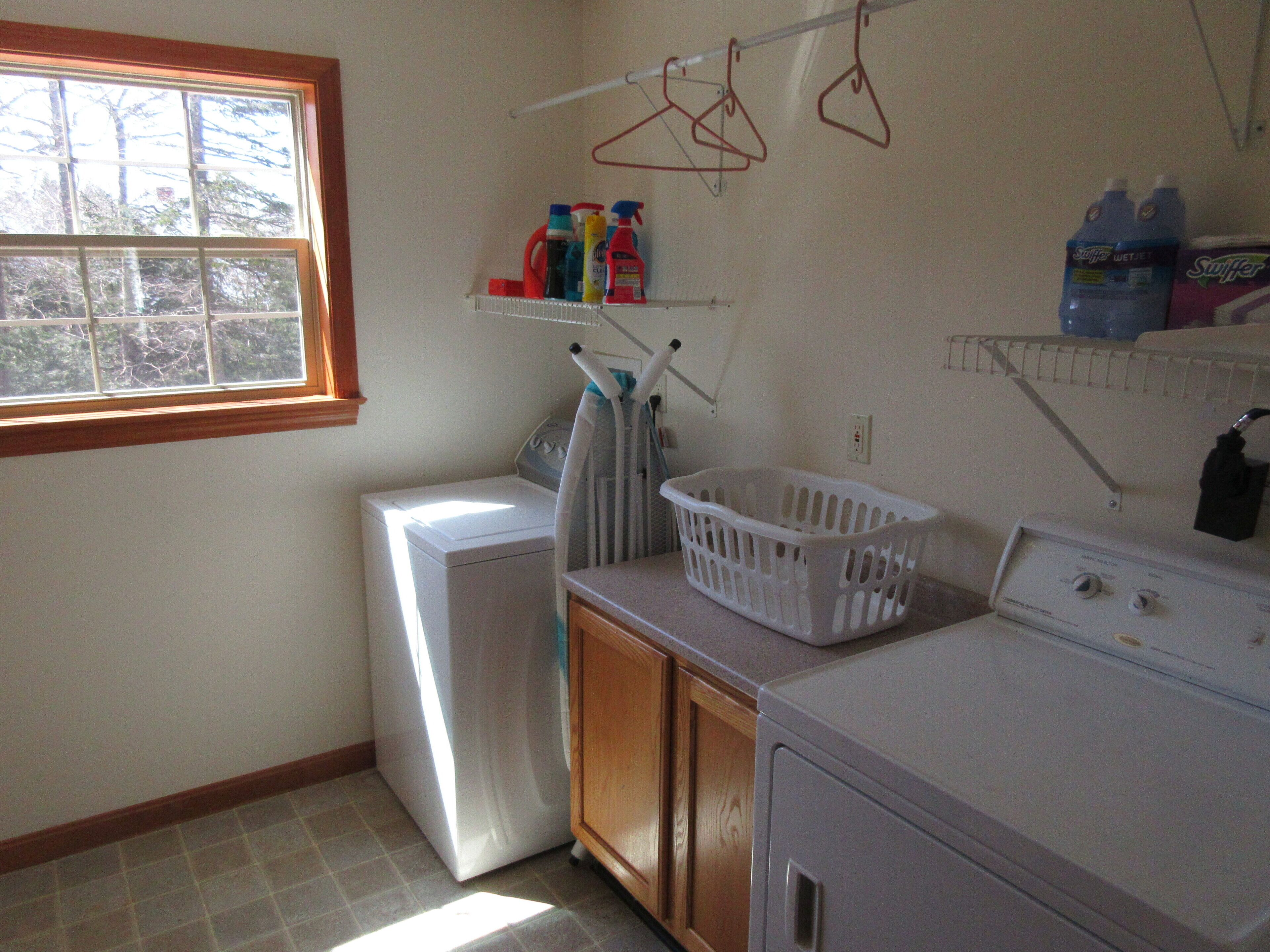 A laundry room with a washer, dryer, and ironing board. There are shelves with cleaning supplies and a window with a view of trees.