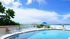 A clear blue pool with lounge chairs and umbrellas, surrounded by greenery and a view of the ocean. - Beachcomber unit 501 (Longboat Key)