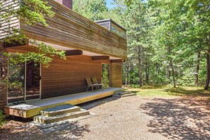 A modern wooden cabin with a covered porch, built-in seating, and a gravel driveway surrounded by trees. - Architectural modern home 5 mins to beach! (Truro)