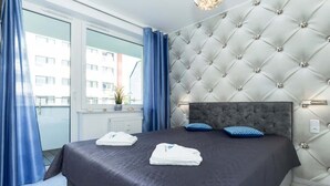 A hotel room with a large bed, a tufted headboard, a window with curtains, and a view of buildings outside. - Stylish Apartment with Gym | 400 m to the Beach, Balcony (Kolobrzeg)