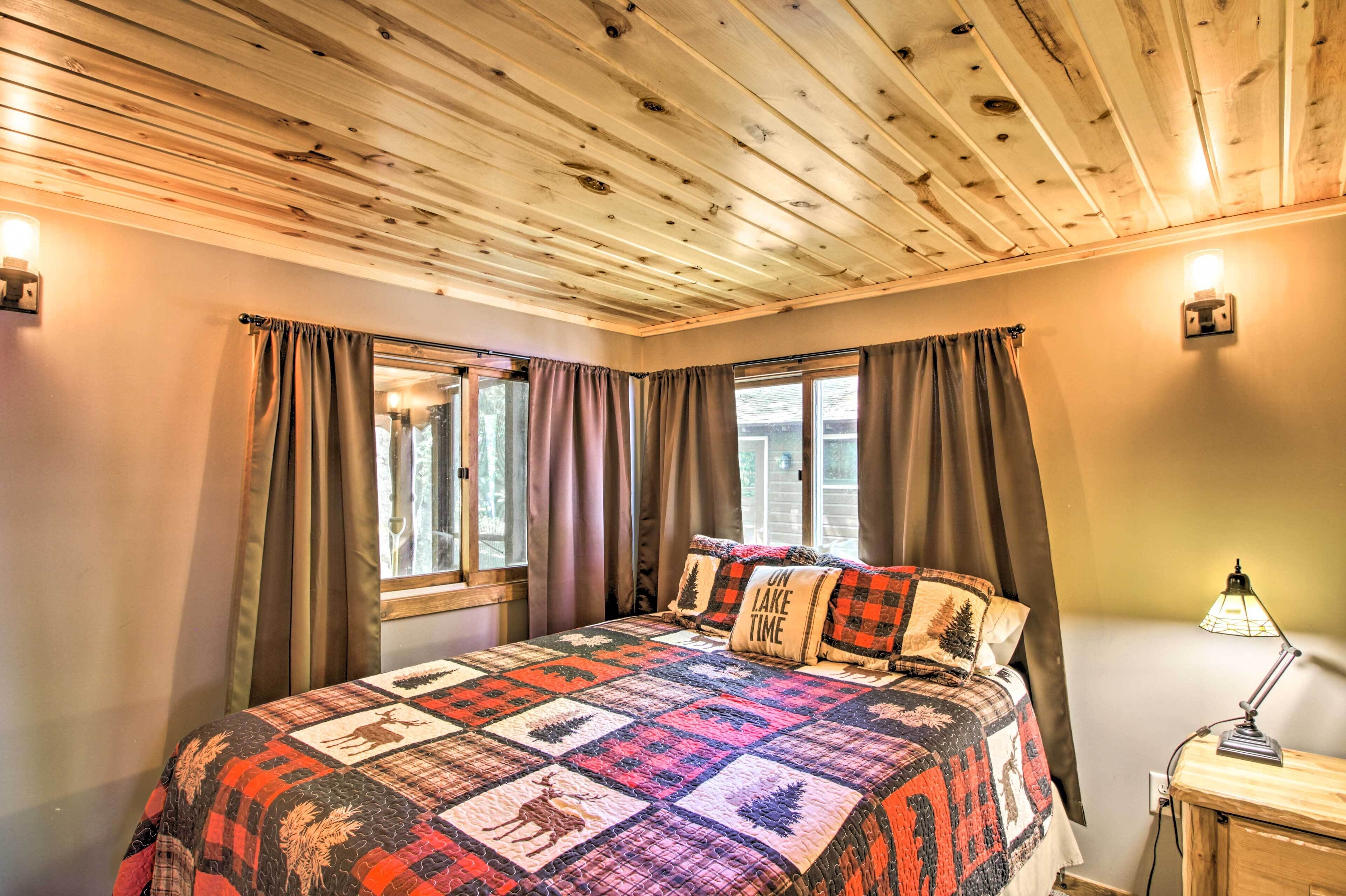 A bedroom with a wooden ceiling, a quilted bedspread, two windows with curtains, and a bedside lamp.