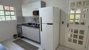 Una cocina con gabinetes blancos, microondas, refrigerador, ventilador de techo y una ventana con persianas.
