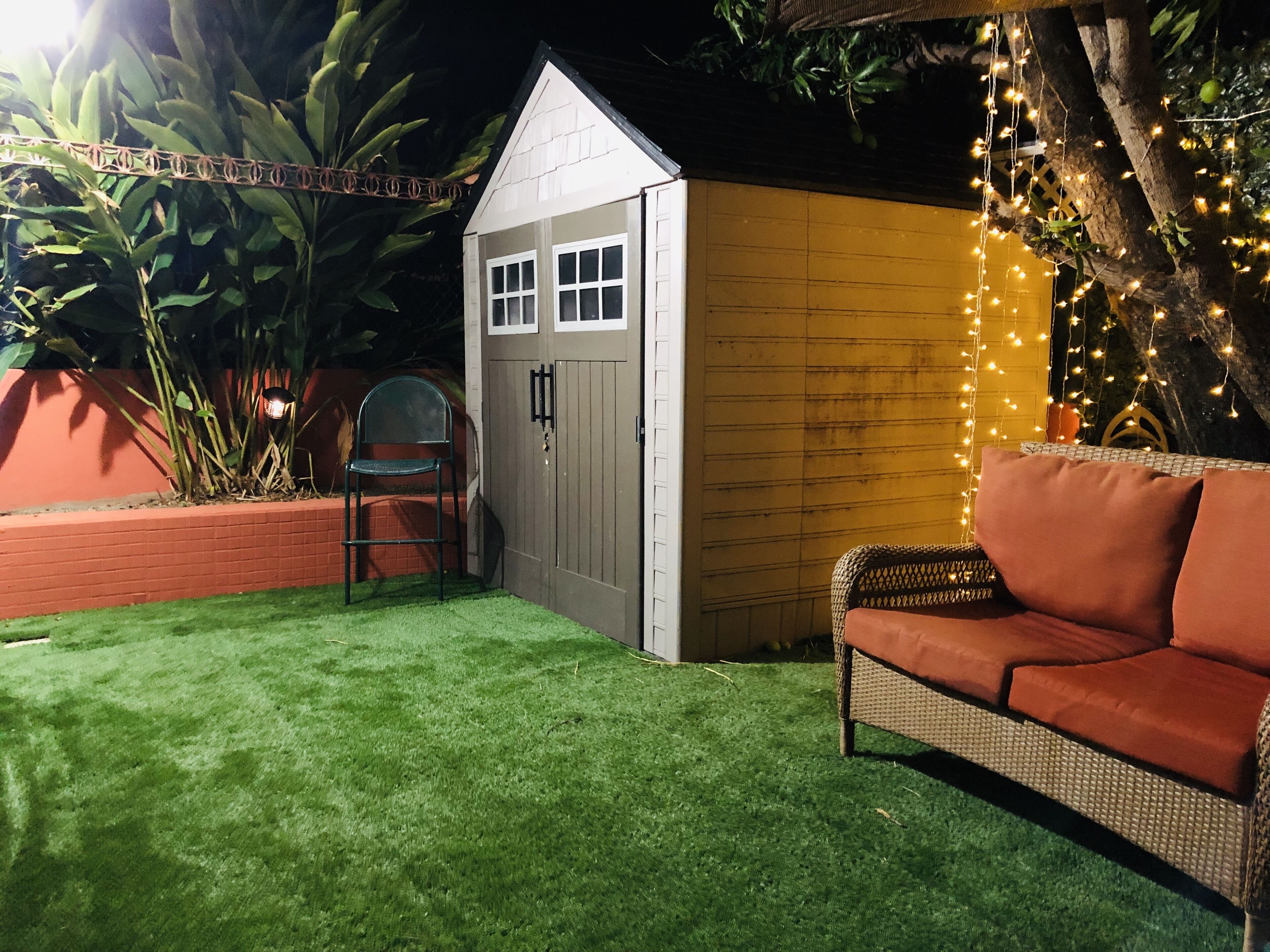 Un petit abri de jardin avec une porte et des fenêtres, une pelouse artificielle verte, un canapé en osier et des guirlandes lumineuses.