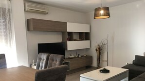 Una sala de estar moderna con una mesa de comedor de madera, sillas tapizadas en gris, una mesa de centro blanca, un sofá gris, un televisor de pantalla plana, un estante empotrado y una lámpara colgante.