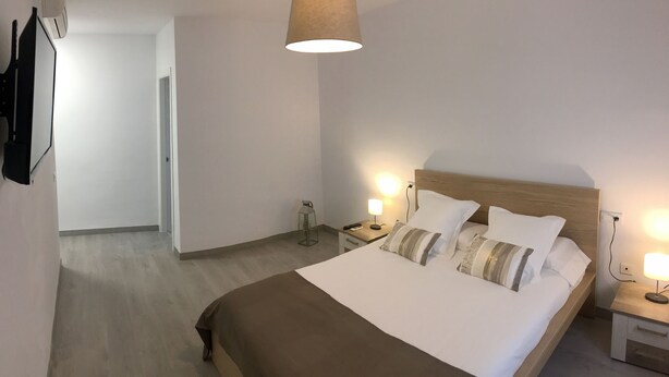 Habitación de hotel con cama, lámparas de noche, cabecero de madera, mesita de noche, televisor montado en la pared y una puerta que da a otra habitación.