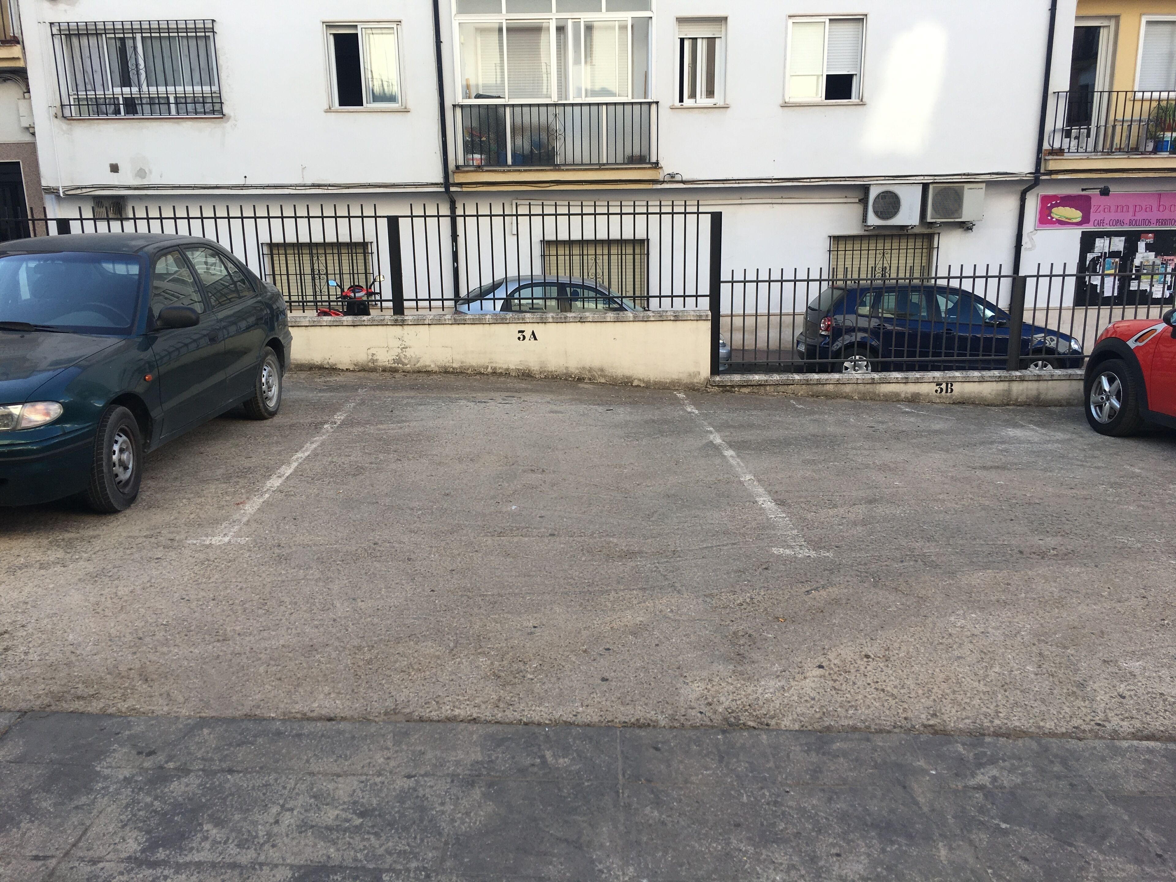 Zona d'aparcament amb un edifici blanc al fons, una tanca negra i diversos cotxes estacionats.