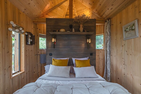 A wooden cabin bedroom with a bed, two wall-mounted light fixtures, a shelf with decorative items, and a window with curtains. - Treehouse 2 people - spa & pool - exceptional view (Montréal-les-Sources)