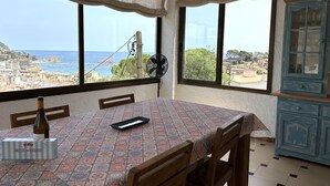 A dining area with a table set for two, a bottle of wine, and a box of crackers. There are two windows with views of a coastal town and a wind turbine. - BEAUTIFUL HOUSE SEA VIEWS (Tossa de Mar)