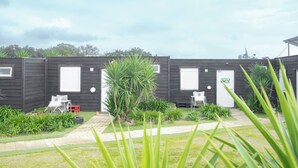 A row of dark-colored mobile homes with white doors and windows, each with a small garden area and a white chair outside. - Bungalow (Montemor-o-Novo)