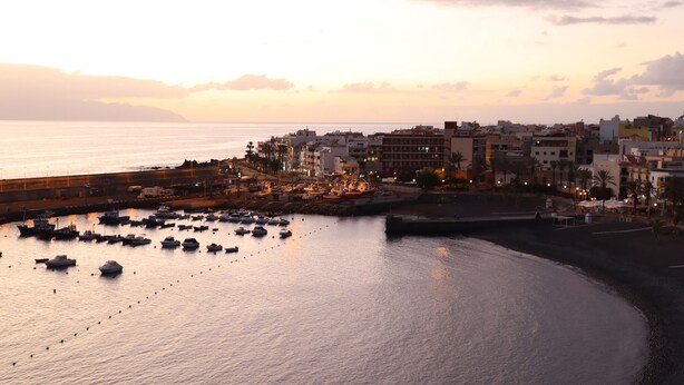 Una cittadina costiera al tramonto con barche ormeggiate nel porto, edifici e una spiaggia sabbiosa.