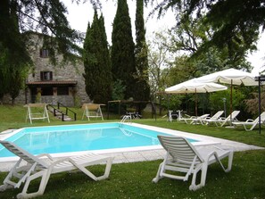 Una piscina con sillones blancos y sombrillas, rodeada de vegetación y un edificio de piedra.