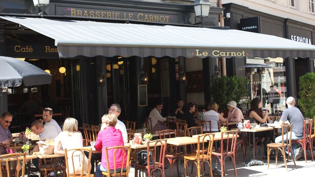 Un café al aire libre con mesas y sillas, un letrero que dice "Le Carnot", y una fachada con "Brasserie Le Carnot" en la parte superior.