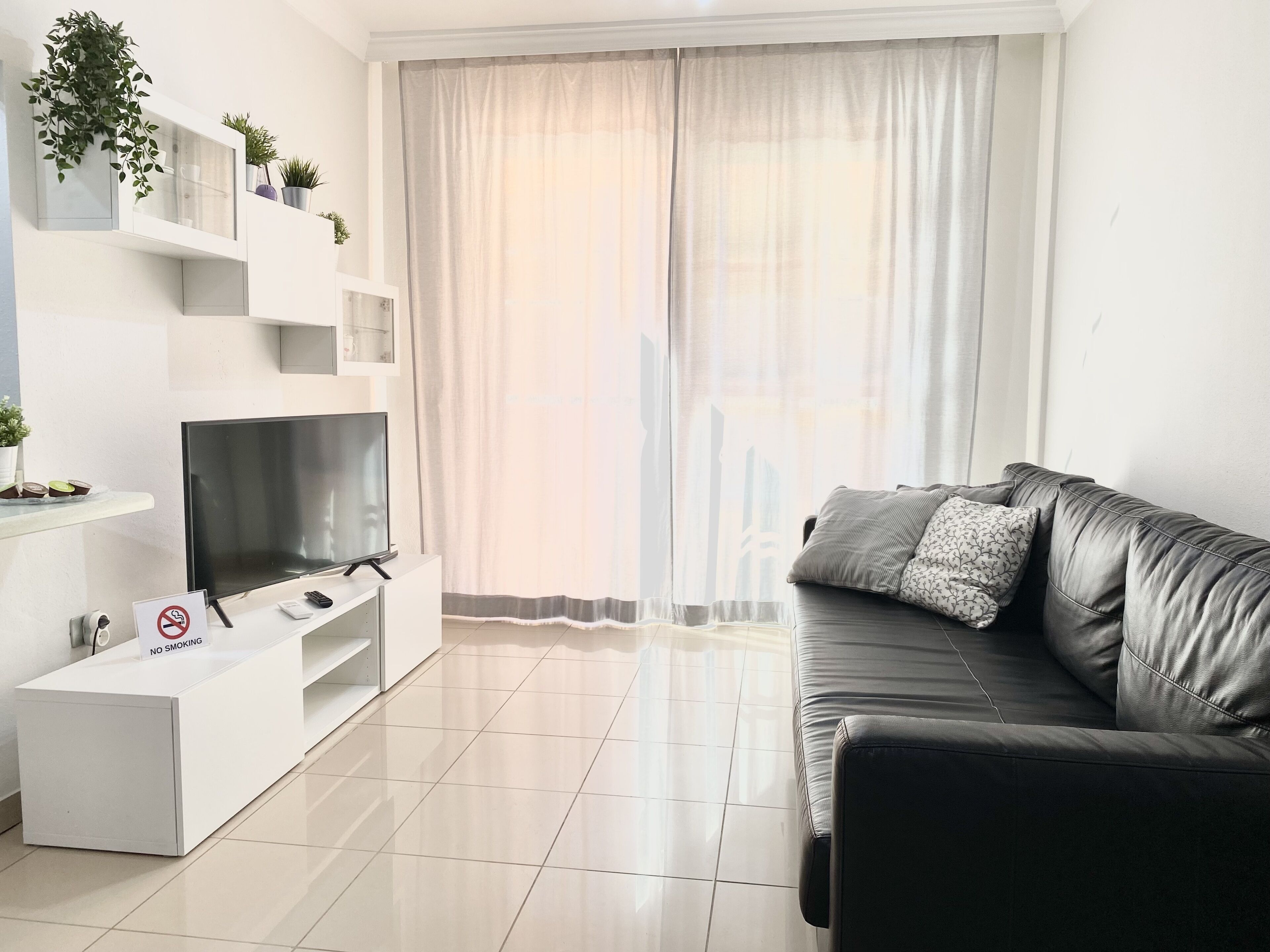 A modern living room with a flat-screen TV, a white entertainment unit, a leather sofa, and a large window with sheer curtains.