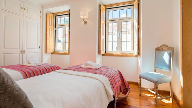 A bedroom with two beds, a wooden chair, and a window with a view of buildings. - Gomes Freire Terrace (Cascais)
