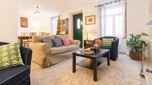 A living room with a sofa, armchair, and coffee table. There is a dining table with chairs, a lamp, and a green door. - Gomes Freire Terrace (Cascais)