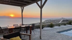 Un patio con una mesa y sillas, con vista a una piscina y una vista panorámica del mar y montañas distantes durante el atardecer.