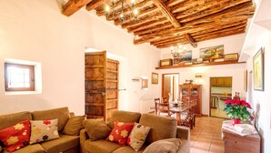 Un salon chaleureux avec un plafond en bois, un ensemble de canapés, un coin repas et une cuisine visible à travers une porte ouverte.