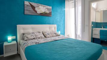 Ein Schlafzimmer mit einer türkisblauen Bettdecke, einem weißen Kopfstück und einem Nachttisch mit einer türkis-farbenen Lampe. Über dem Bett hängt ein Bild mit weißen Blumen, und rechts ist ein Badezimmer mit einem blauen Waschbecken und Spiegel zu sehen.