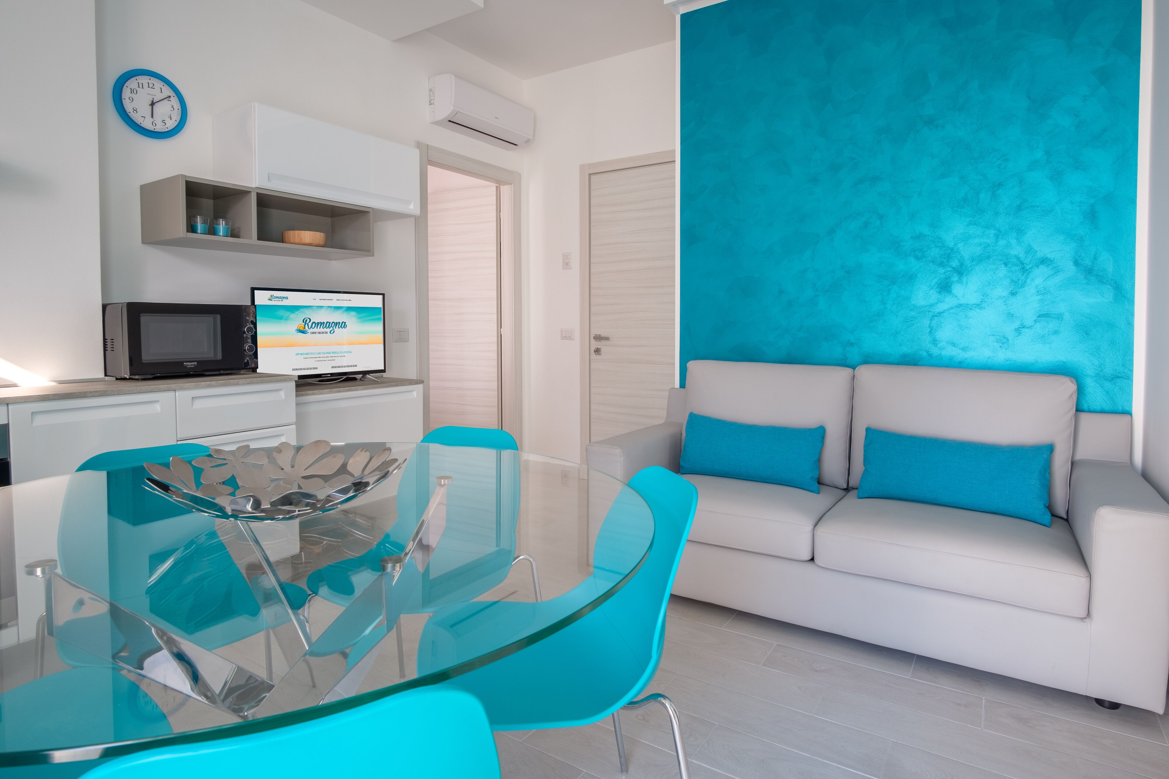 Un soggiorno moderno con un tavolo da pranzo in vetro, un divano bianco con cuscini blu e un angolo cottura con forno a microonde e una sveglia a parete.