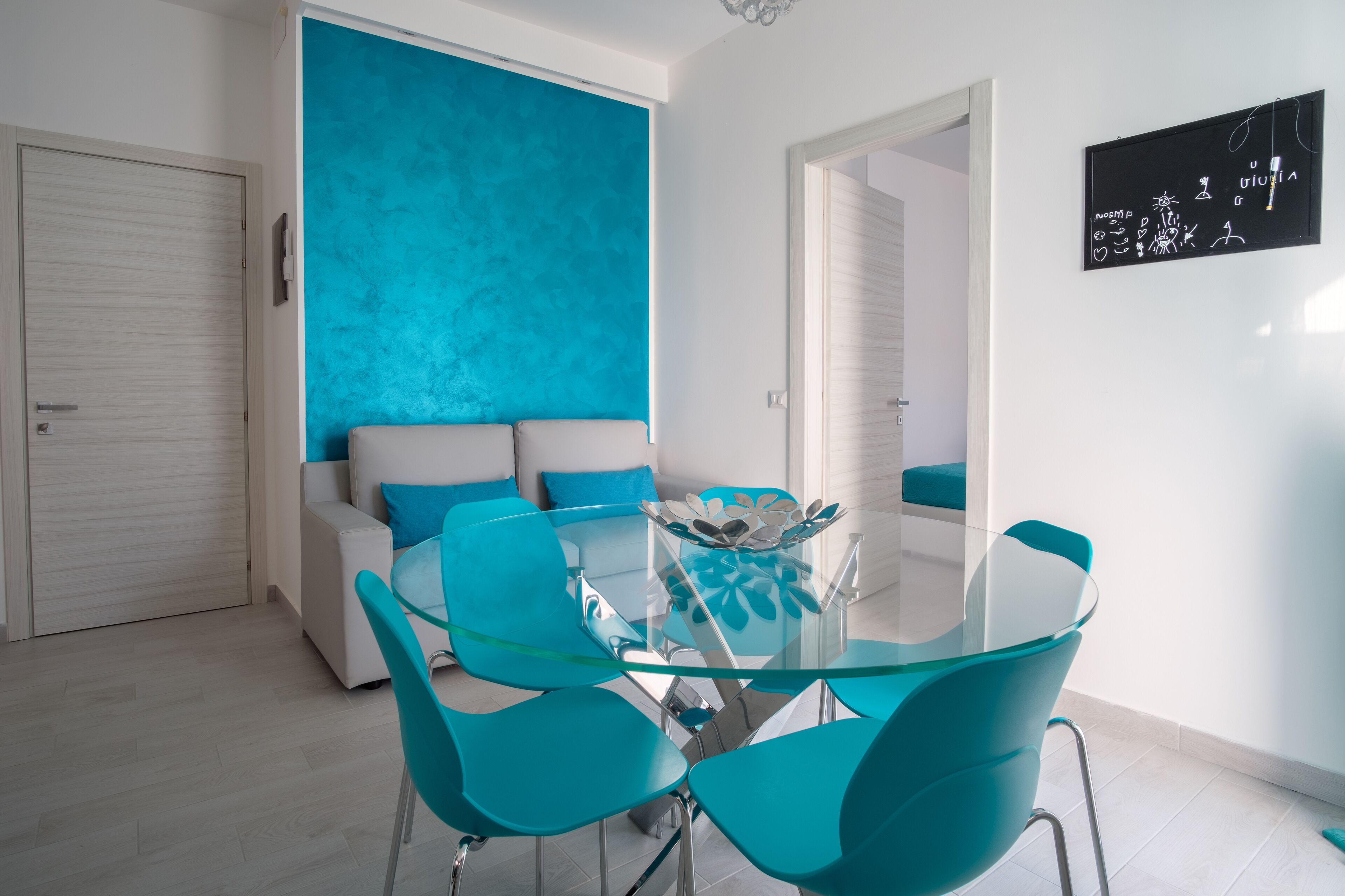A modern dining area with a glass table and teal chairs, a sofa with blue cushions, and a wall with a chalkboard.