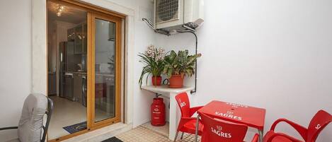 Ein kleiner, umzäunter Außenbereich mit einer Glastür zu einer Küche, einem Tisch mit roten Stühlen und einem Blumentopf auf einem weißen Tisch.