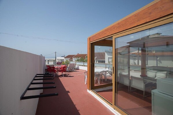 A rooftop terrace with a red floor, a wooden wall, and a glass door leading to a seating area with outdoor furniture.