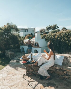 A stone patio with a table set for a meal, a person in a white dress, and a man in a hat. - On The Rocks - The Main House - Serviced Villa (Ios)