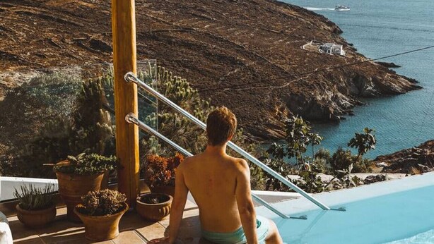 A man sitting by a pool with a view of the sea and rocky terrain.