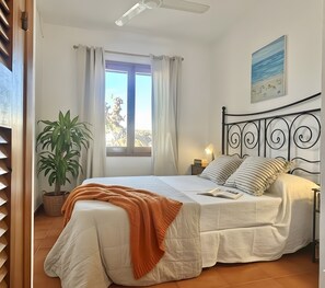 A bedroom with a bed, a window with curtains, a potted plant, a ceiling fan, and a painting on the wall.
