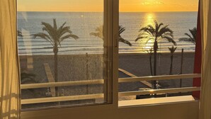 Een kamer met een groot raam met uitzicht op een strand bij zonsondergang.