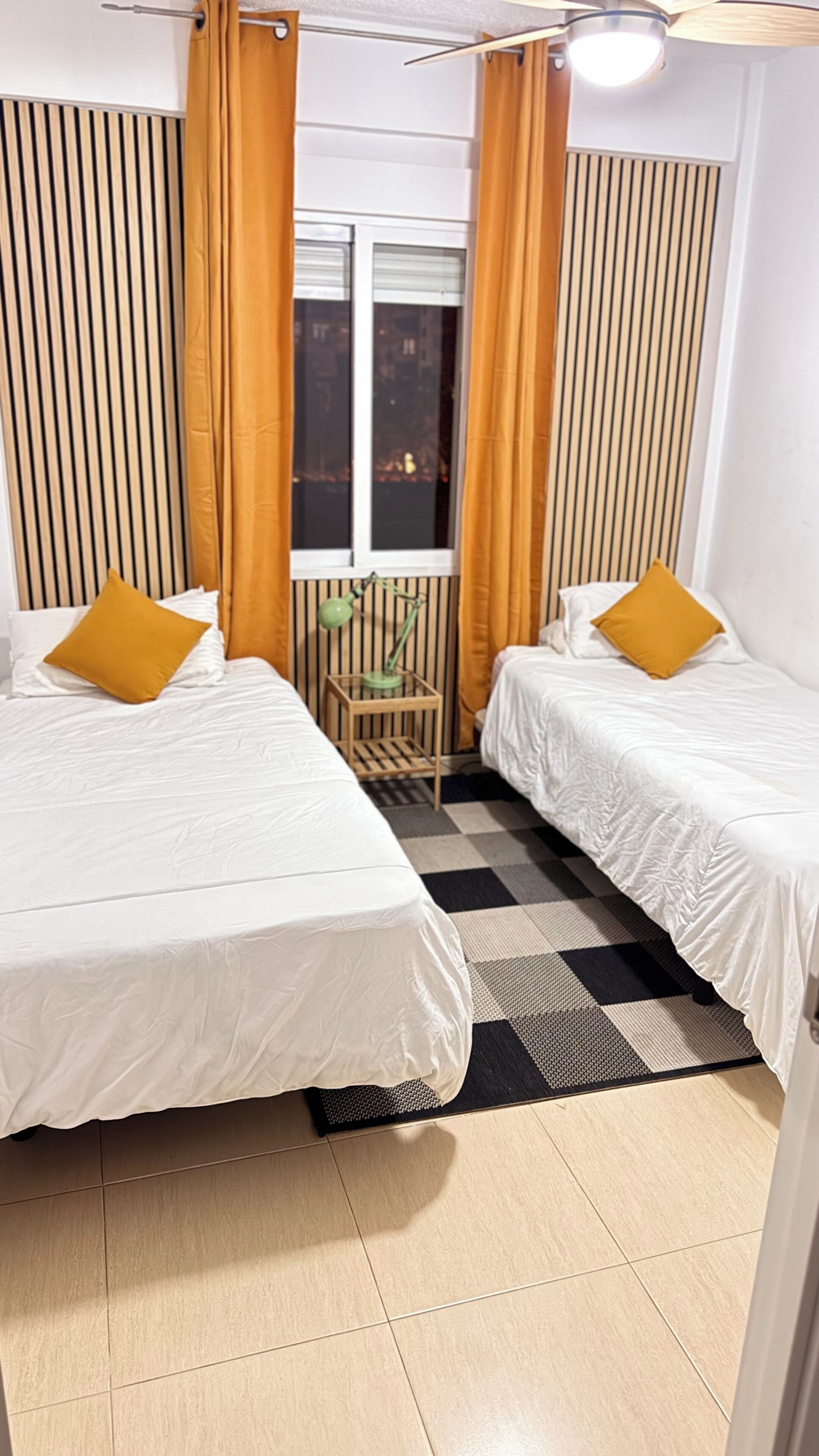 Een kamer met twee bedden, een raam met gordijnen, een klein tafeltje met een lamp en een geruit tapijt.