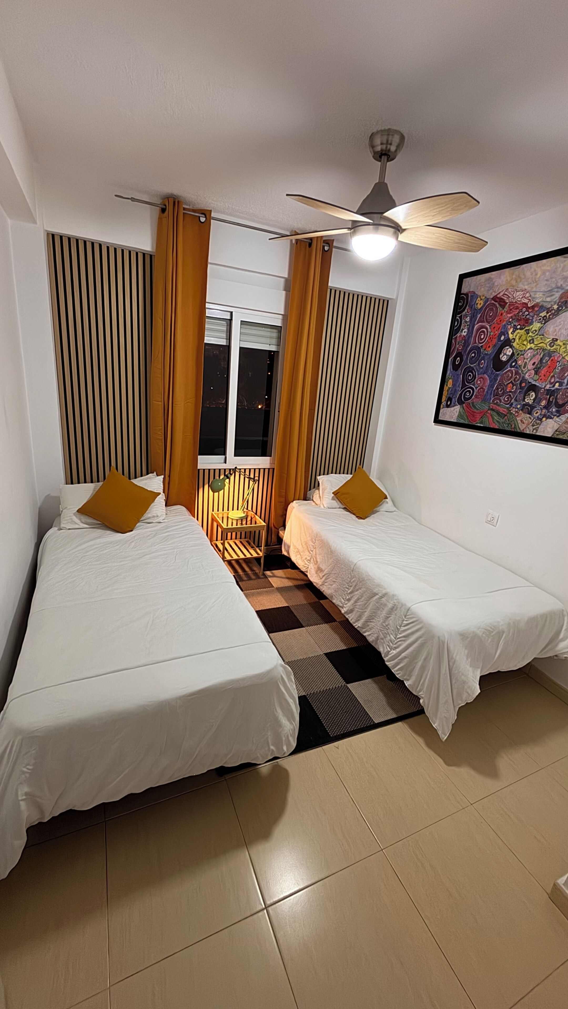 Een kamer met twee bedden, een plafondventilator, een raam met gordijnen, een schilderij aan de muur en een geruit tapijt.