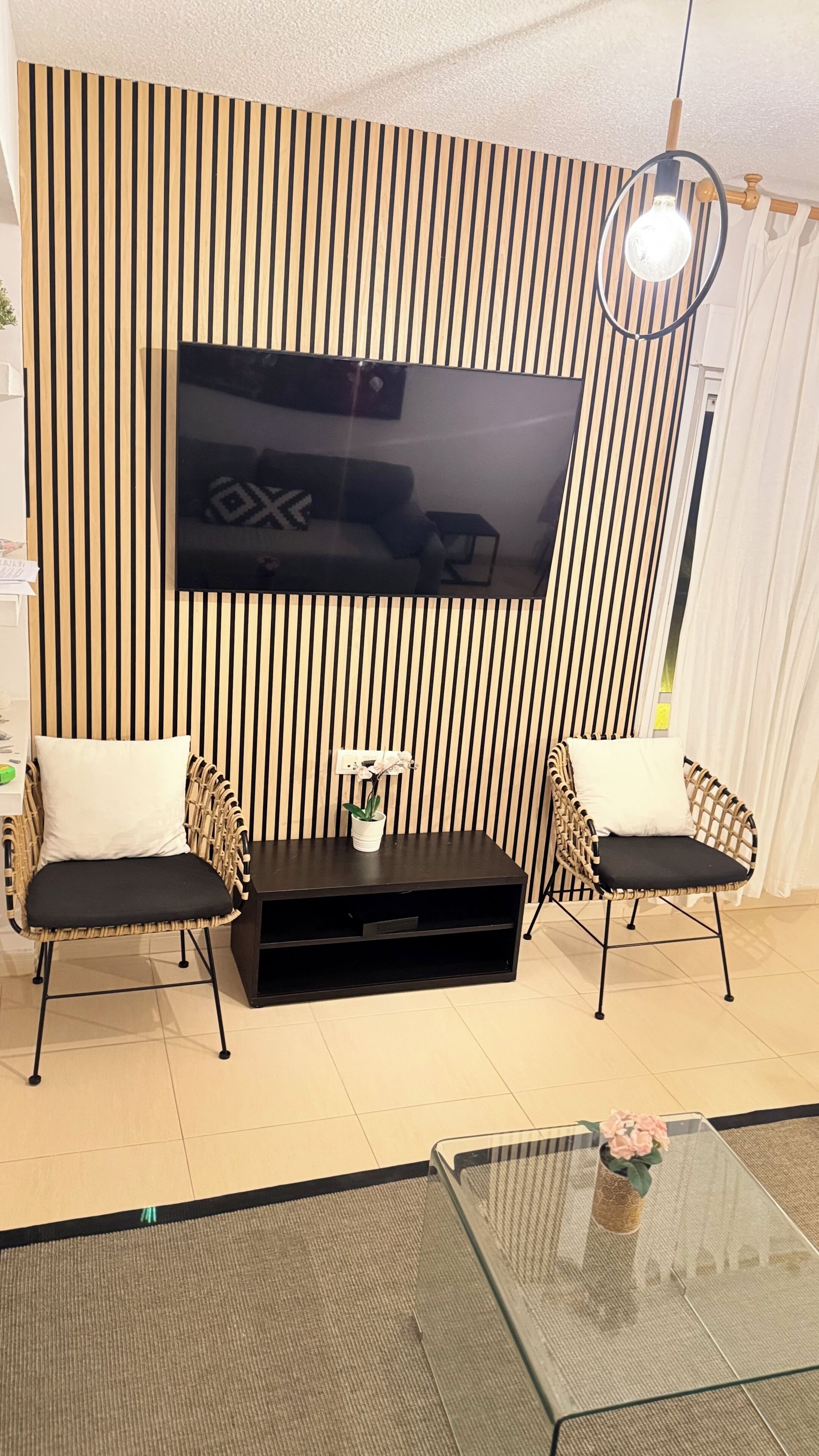 A living room with a flat-screen TV mounted on a striped wall, two wicker chairs with cushions, a glass coffee table, and a small potted plant.