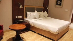 A hotel room with a wooden bed, white bedding, a round wooden table, and a red chair. - The Utsav Grand Banquets & Resort (Sonepat)