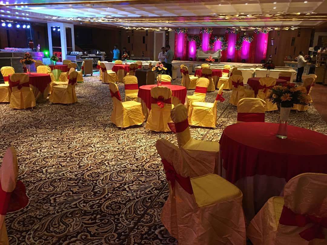 A banquet hall with round tables covered in red tablecloths, chairs with yellow sashes, and a patterned carpet.
