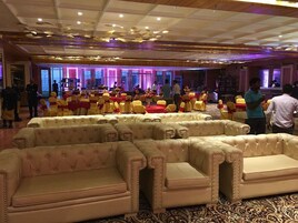Un salón de banquetes con filas de mesas y sillas, un escenario y una zona de estar con sofás.