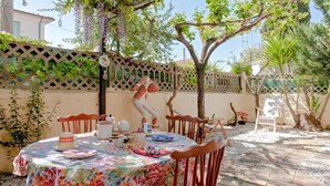 Un tavolo rotondo con tovaglia floreale, circondato da sedie di legno, su un patio in mattoni con un recinto a graticcio e vegetazione sullo sfondo.