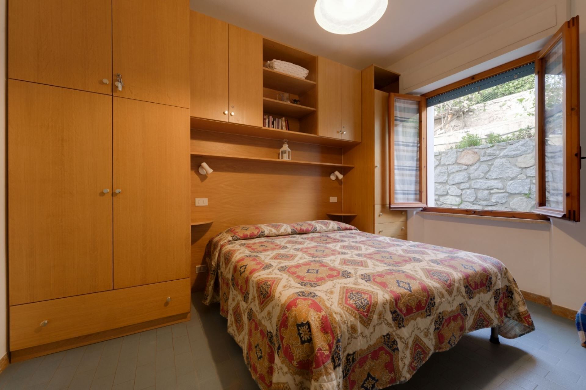 Une chambre à coucher avec un lit, une armoire en bois, des étagères et une fenêtre donnant sur un mur en pierre.