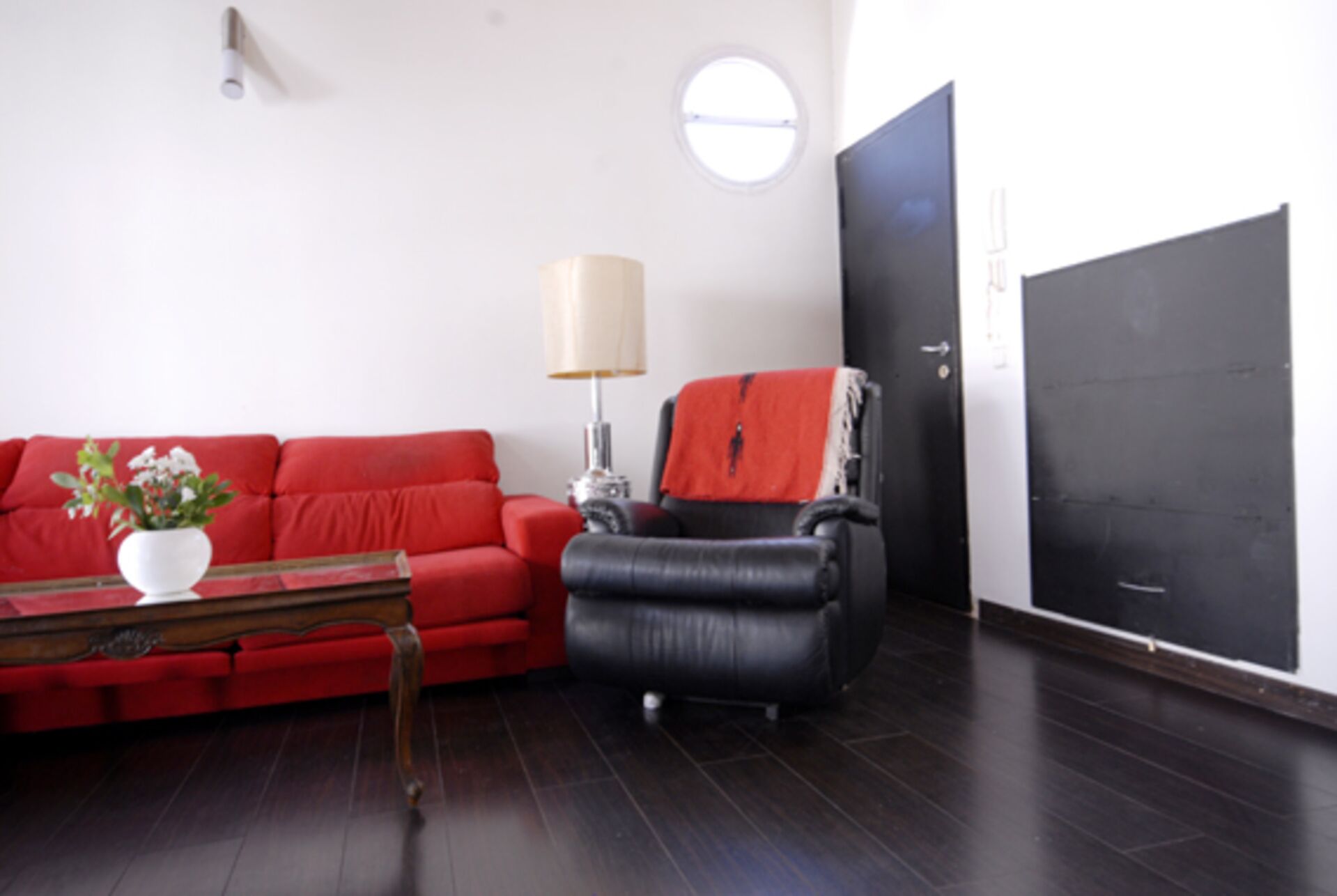 A living room with a red sofa, a black armchair, a small table with a plant, a lamp, and a large black door.