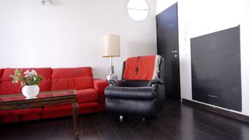 A living room with a red sofa, a black armchair, a small table with a plant, a lamp, and a large black door.