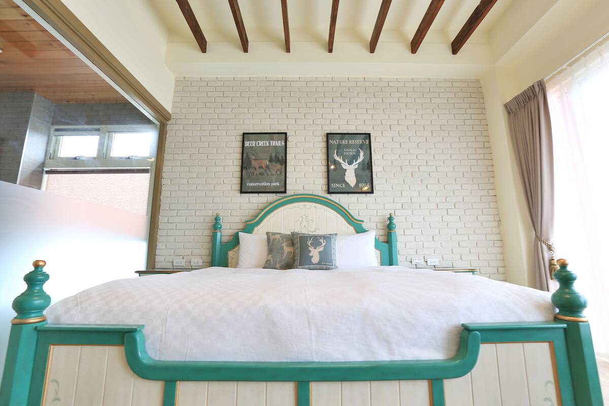 A bed with a white bedspread and teal headboard, two framed pictures on the wall, and a window with curtains.