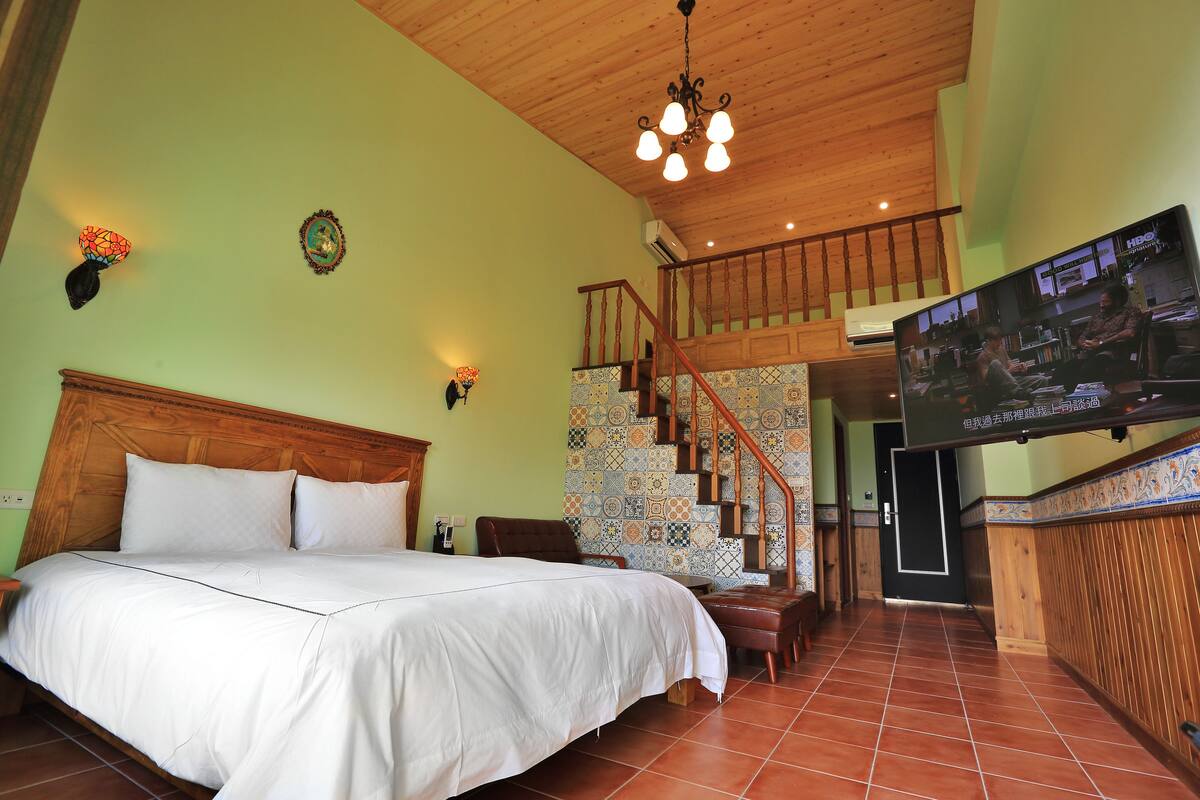 A bedroom with a large bed, a wooden headboard, a staircase, and a TV mounted on the wall.