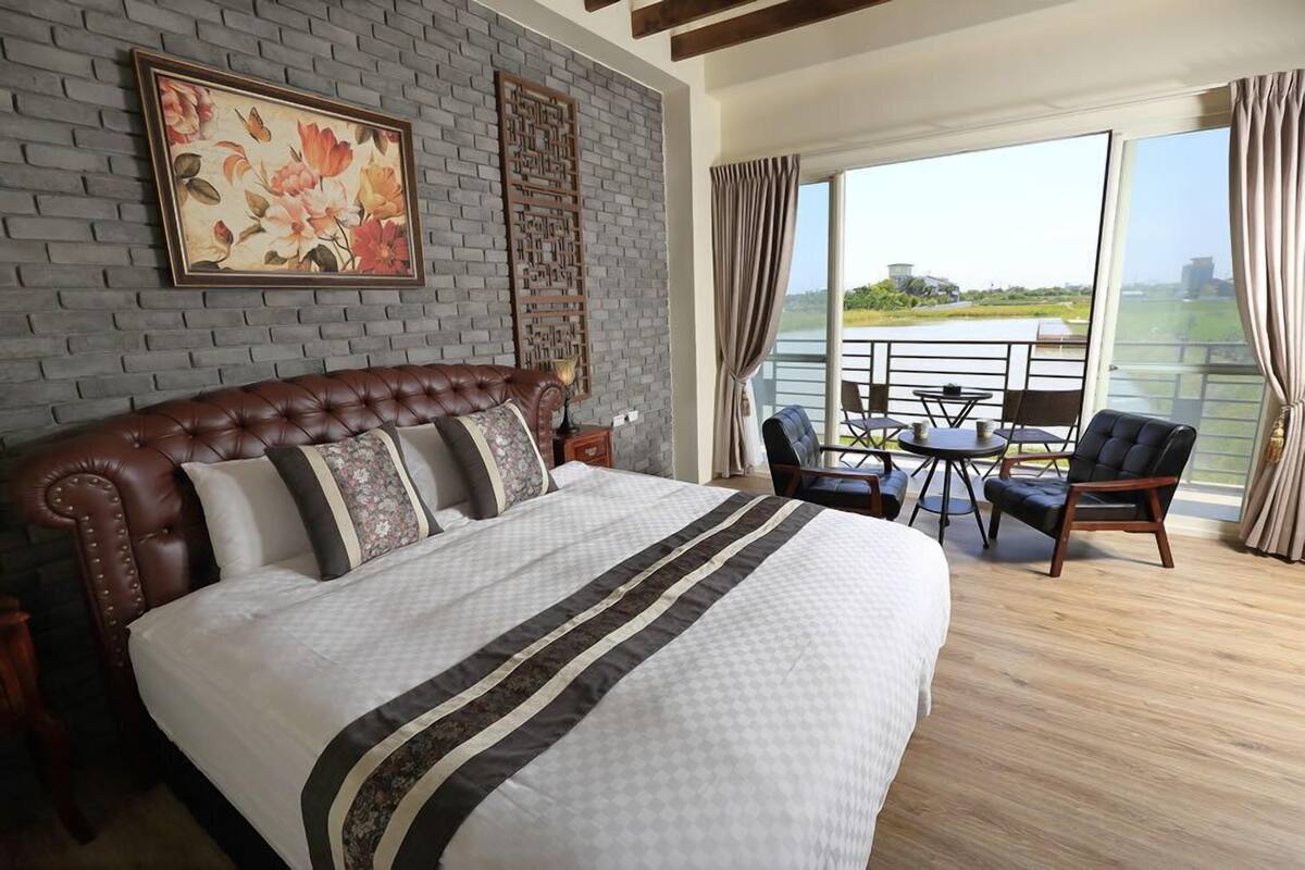 A bedroom with a large bed, a wooden headboard, a decorative wall panel, a framed floral painting, and a balcony with a table and chairs.