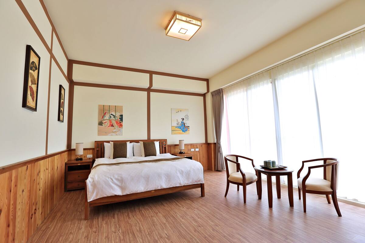 A hotel room with a large bed, wooden headboard, bedside tables, a small round table with chairs, and artwork on the walls.