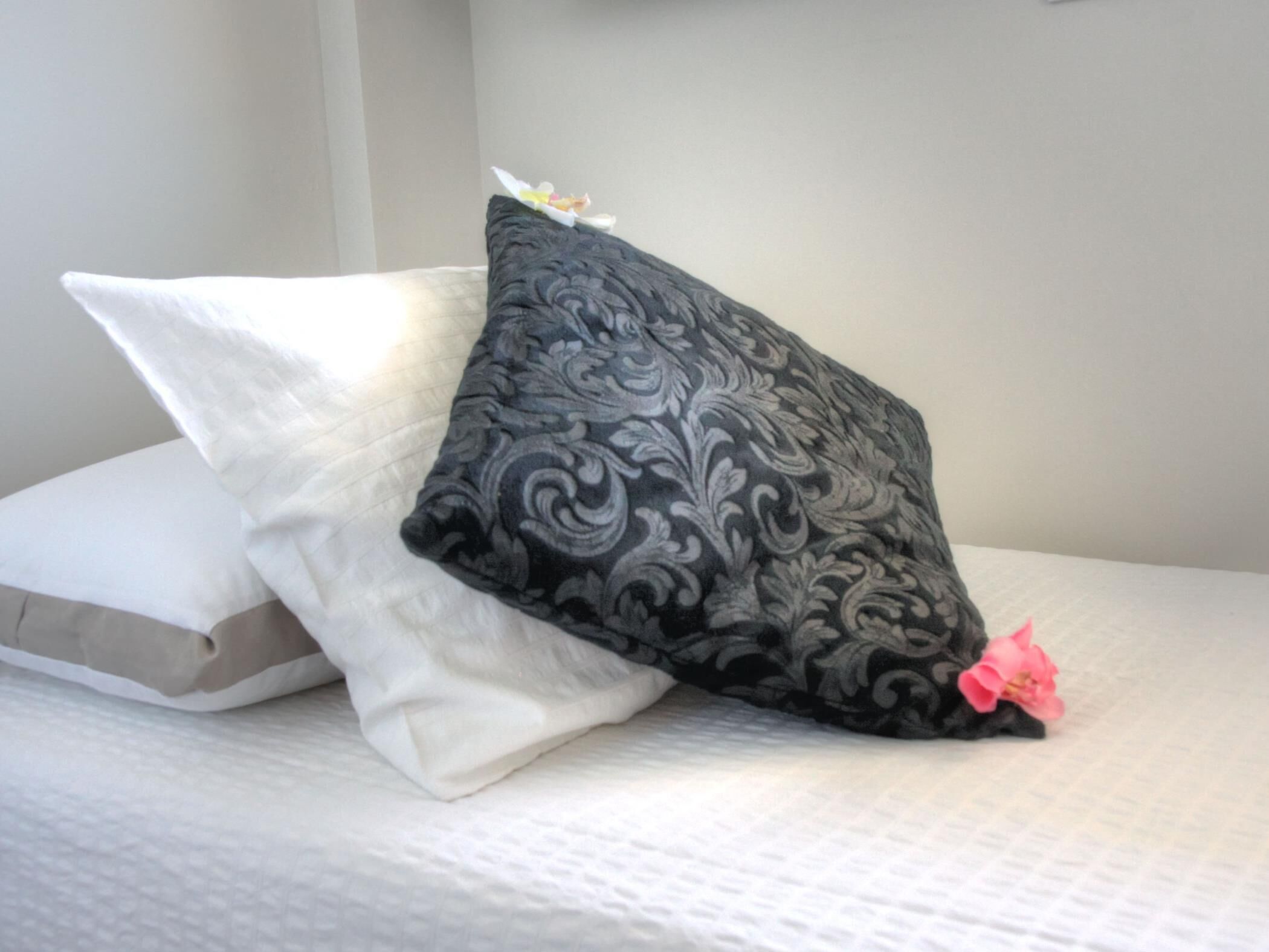 Una cama con tres almohadas: una blanca, una negra con estampado floral y una rosa con una flor.