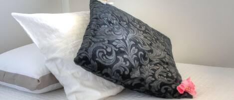 Una cama con tres almohadas: una blanca, una negra con estampado floral y una rosa con una flor.