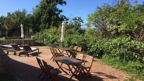 En udendørs terrasse med træborde og stole, omgivet af frodigt grønt og træer.