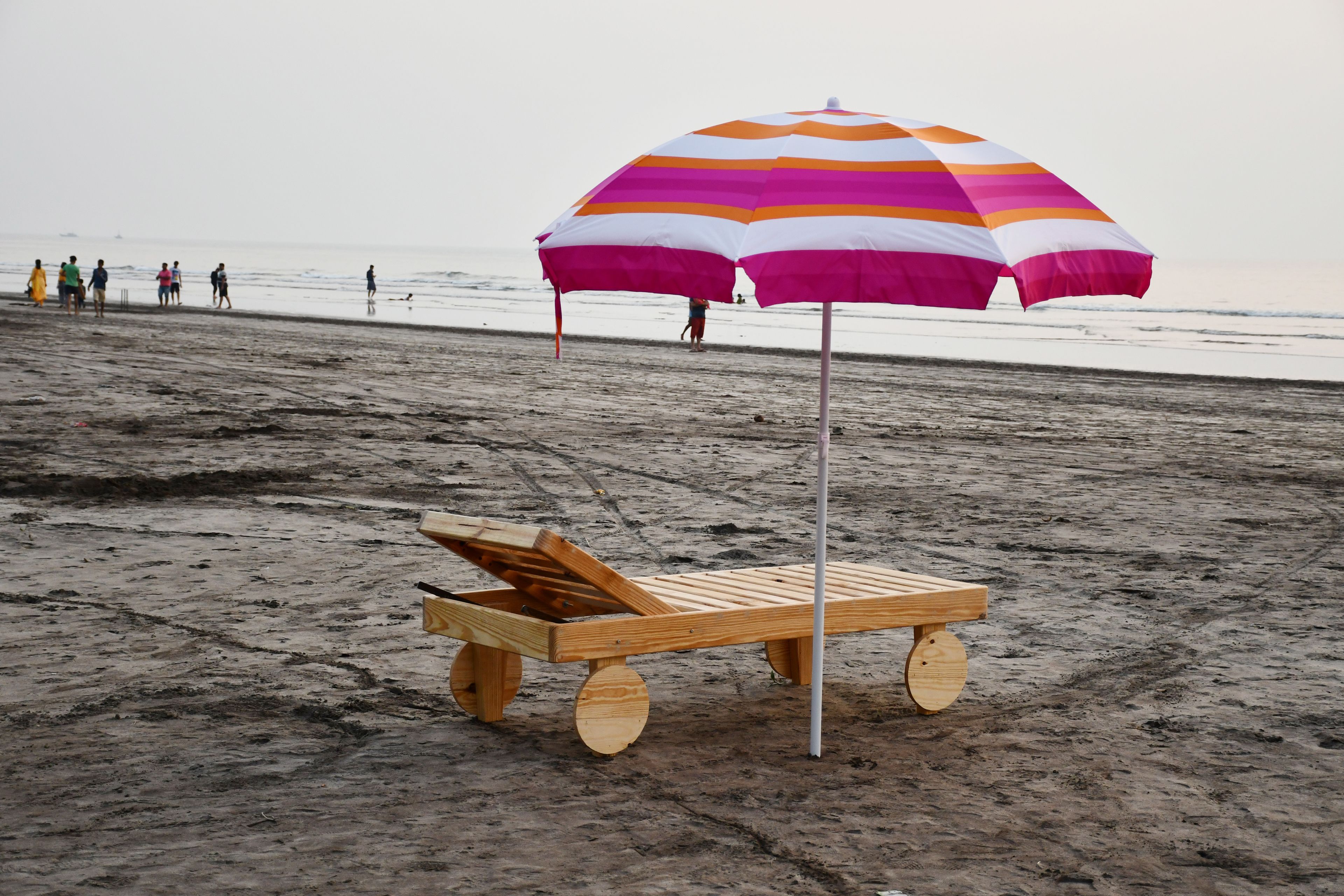 A beach with a wooden lounge chair and a striped beach umbrella.